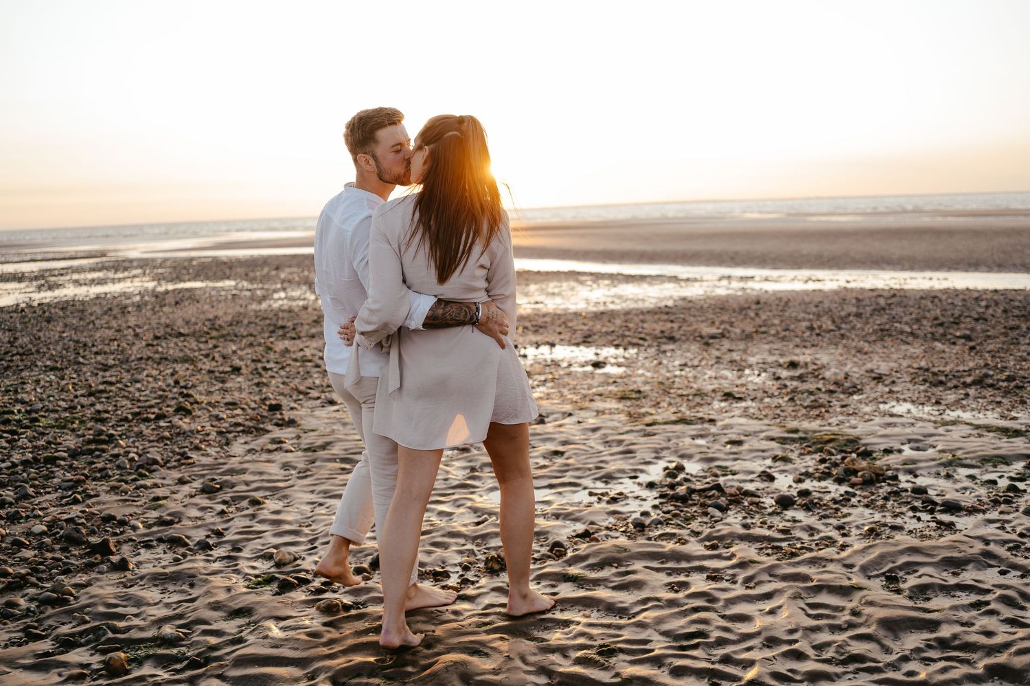 Couple kissing on a sandy beach at sunset with tidal flats in the background.