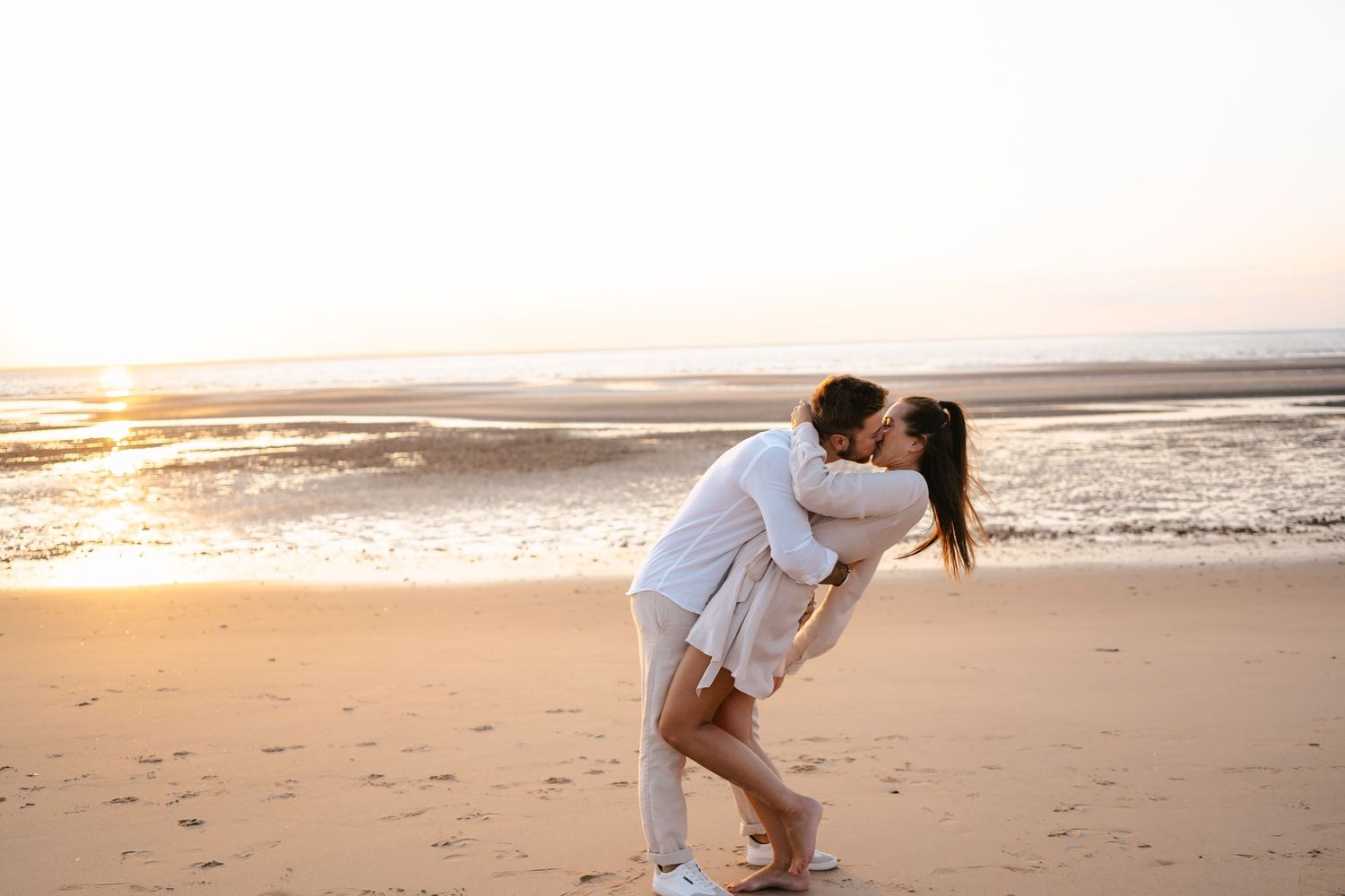 Romantic couple kissing on sandy beach during golden sunset by the ocean.
