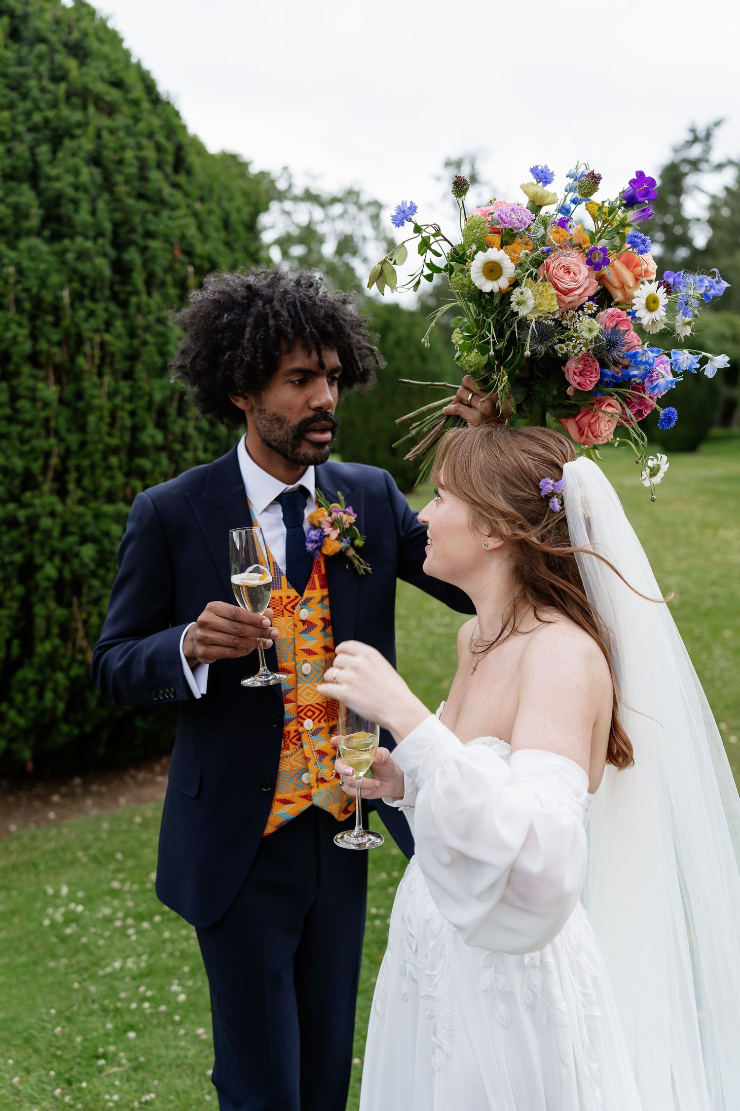 Bride balances large colorful floral arrangement on head while toasting champagne with groom outdoors.