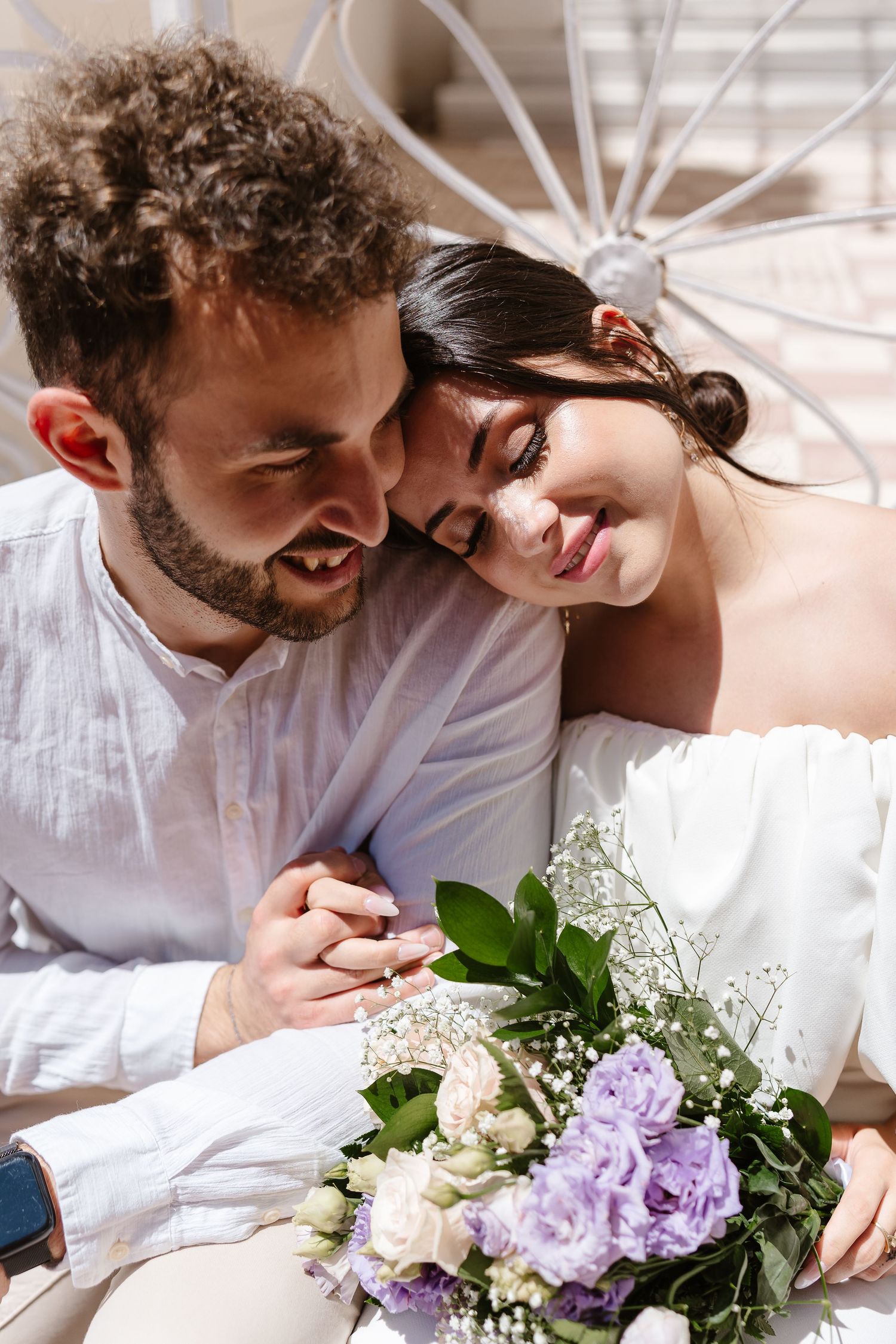 Romantic couple holding hands, bride resting head on groom's shoulder, holding elegant purple and white wedding bouquet.