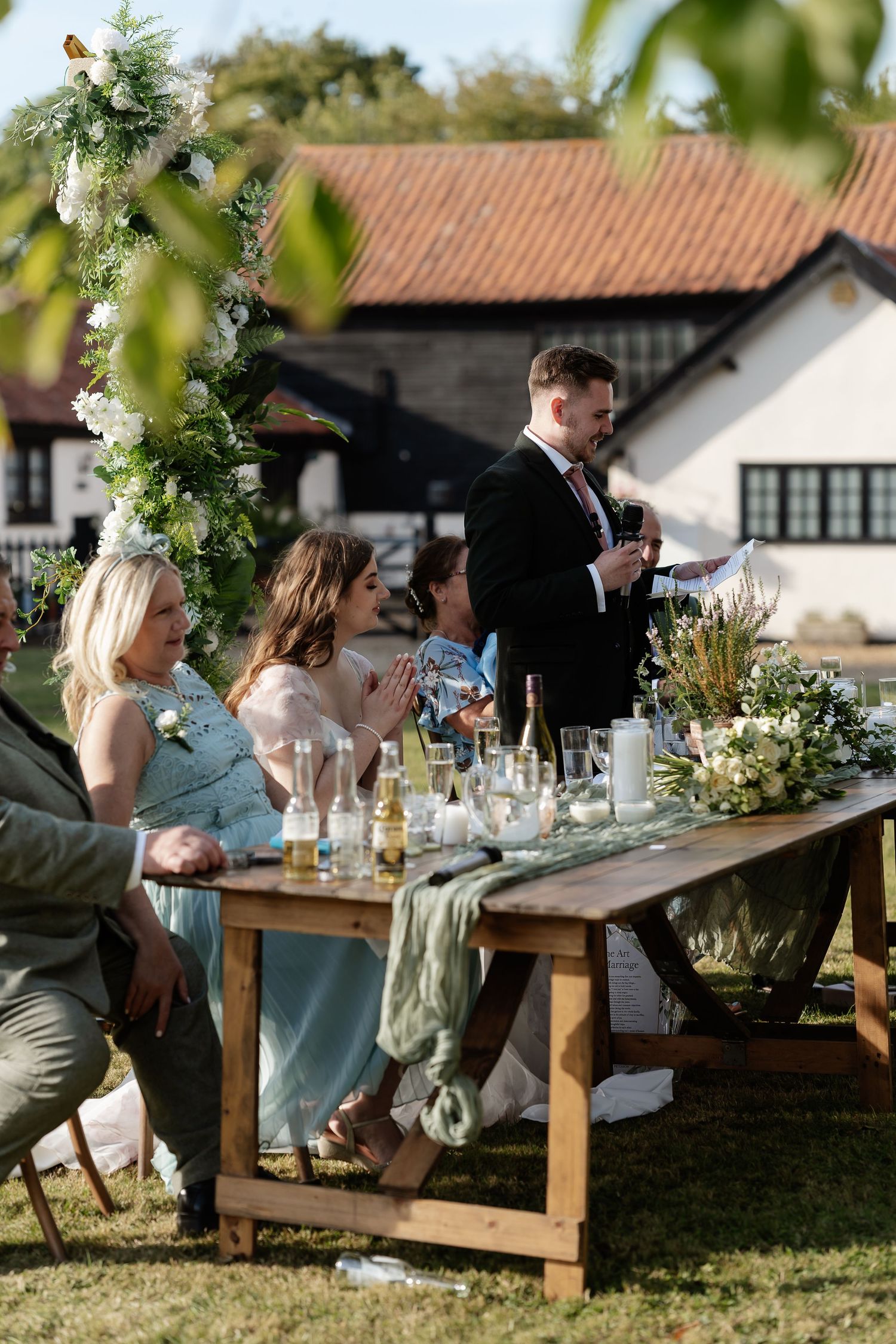 Man giving speech at outdoor wedding reception with guests seated at rustic wooden table and white farmhouse backdrop.