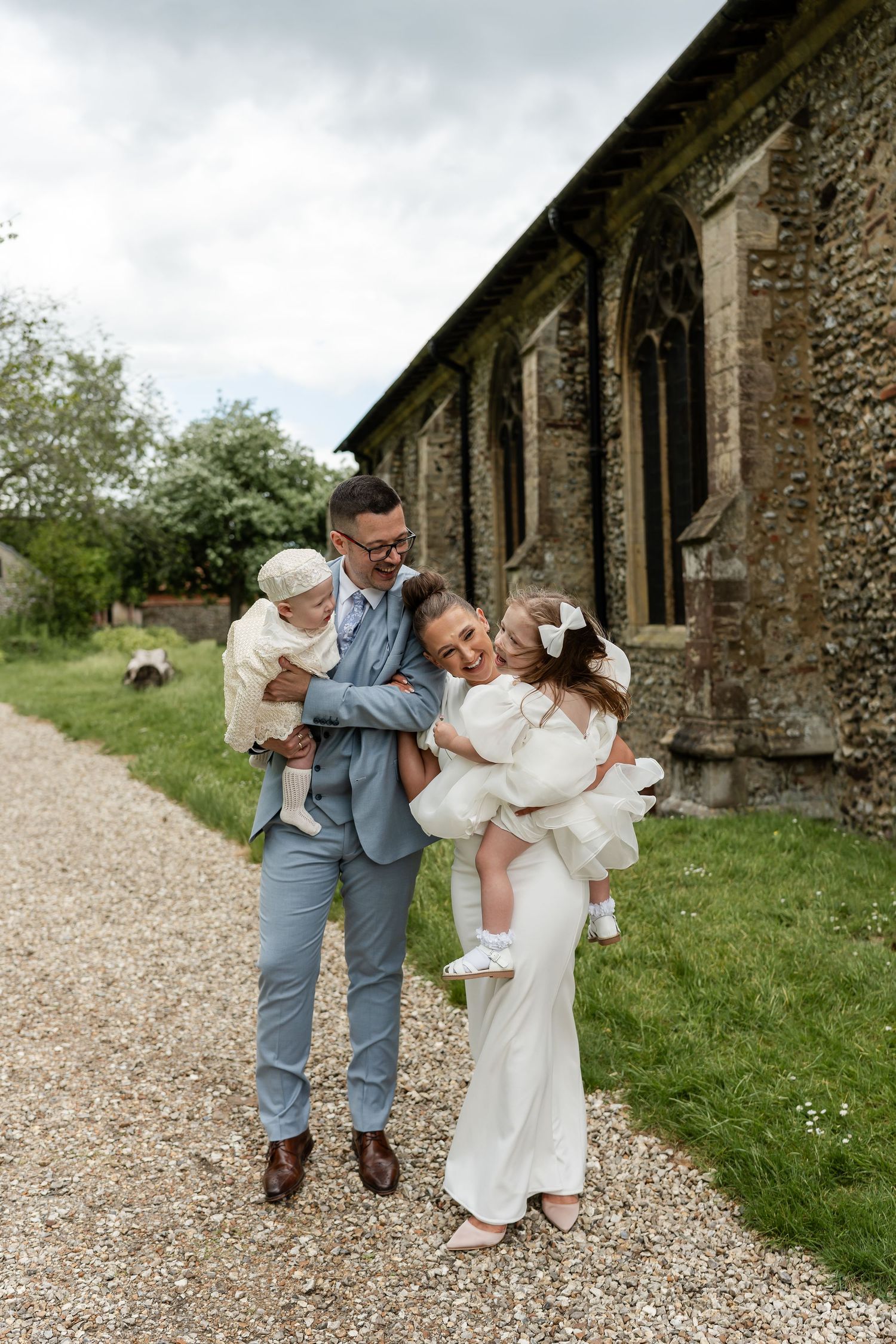 Joyful wedding couple holds two young children outside a traditional stone church on a cloudy day.
