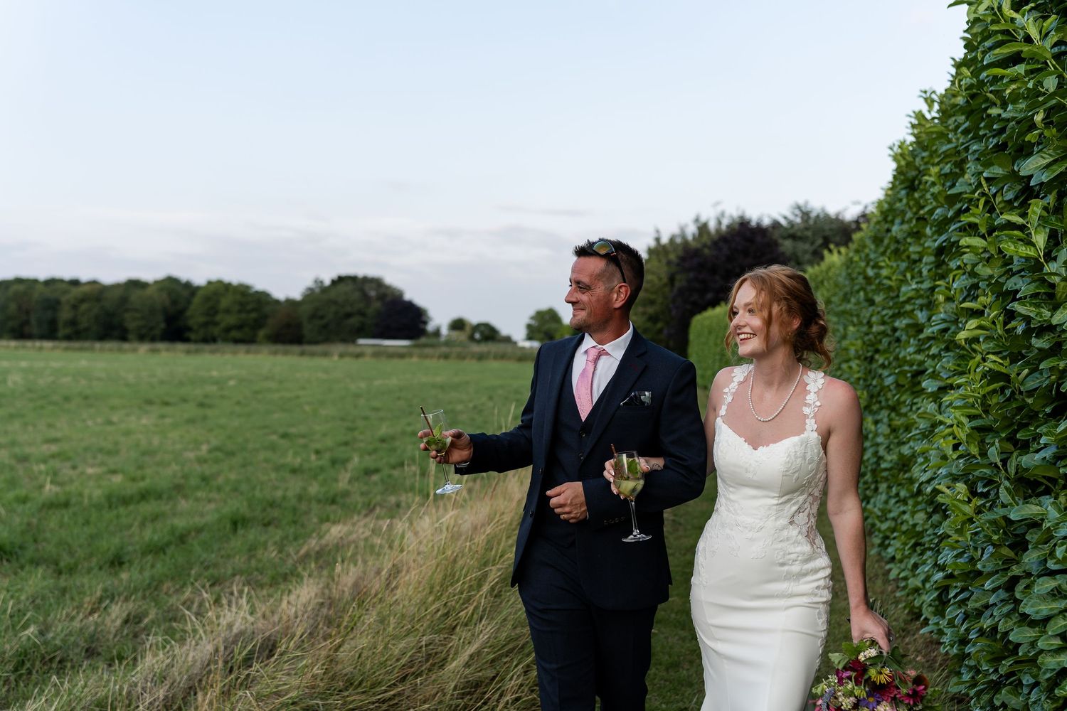 Bride and groom walking hand in hand through green countryside holding champagne glasses.