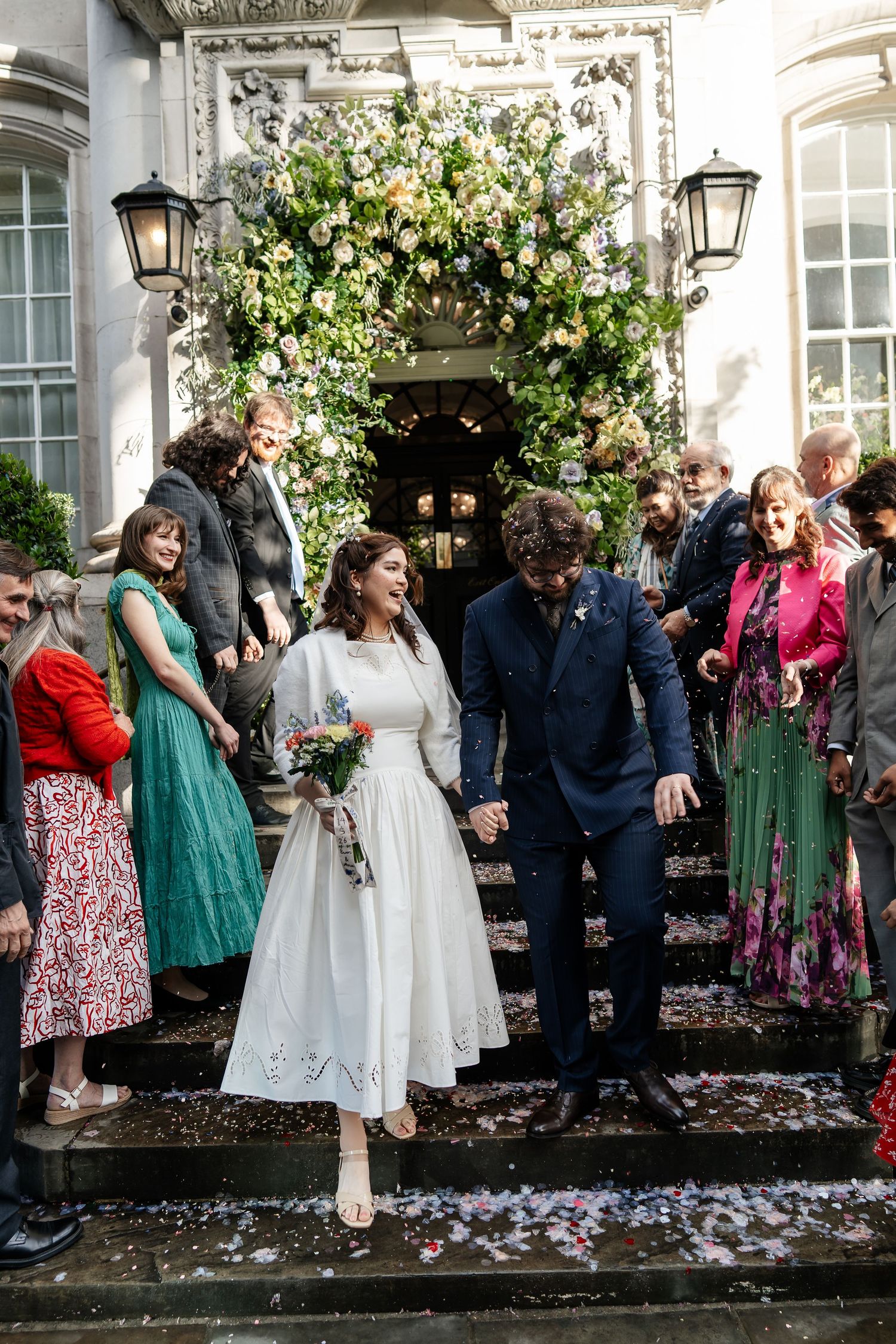 Bride and groom exit venue steps through confetti shower surrounded by smiling wedding guests.