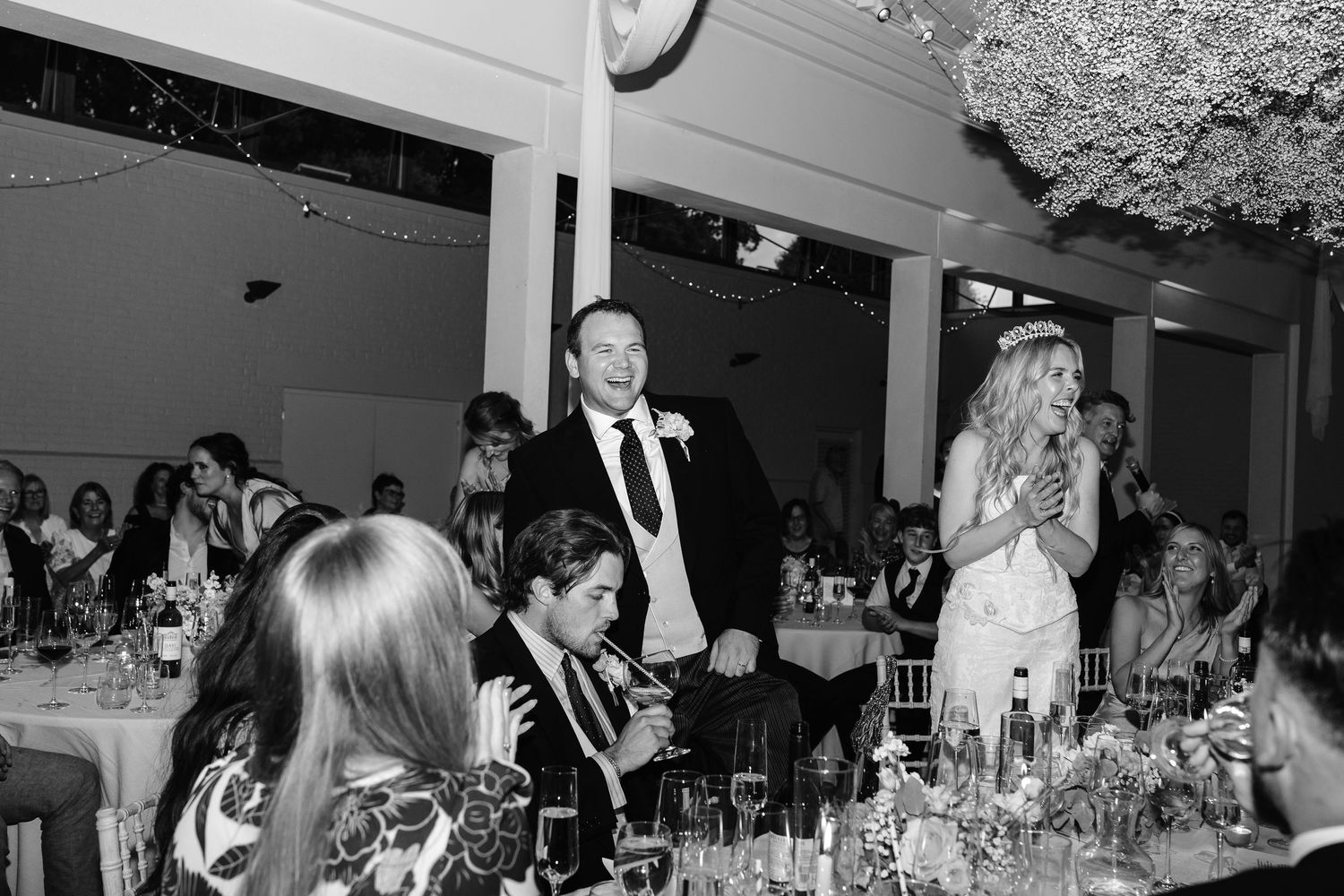 Black and white wedding reception photo of bride giving speech beside groom at elegant floral venue.