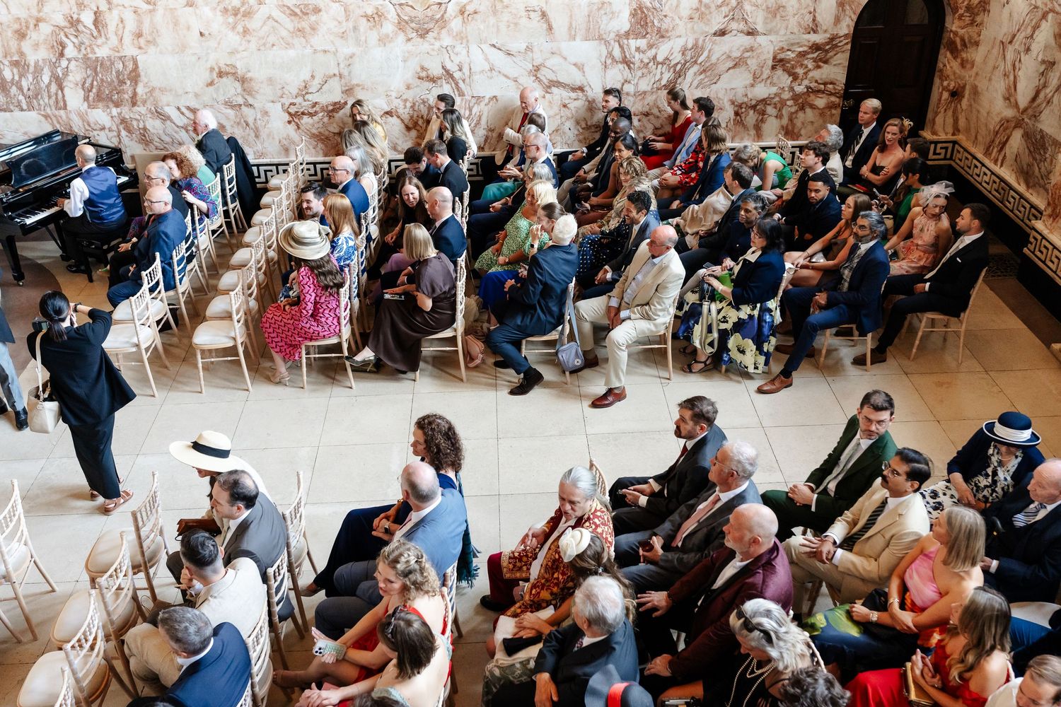 Aerial view of elegantly dressed wedding guests seated in rows inside a grand historic venue.