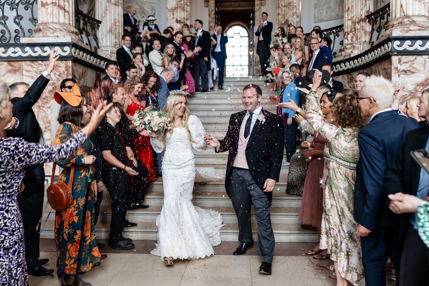 Bride and groom walk through confetti shower on grand staircase surrounded by cheering wedding guests.