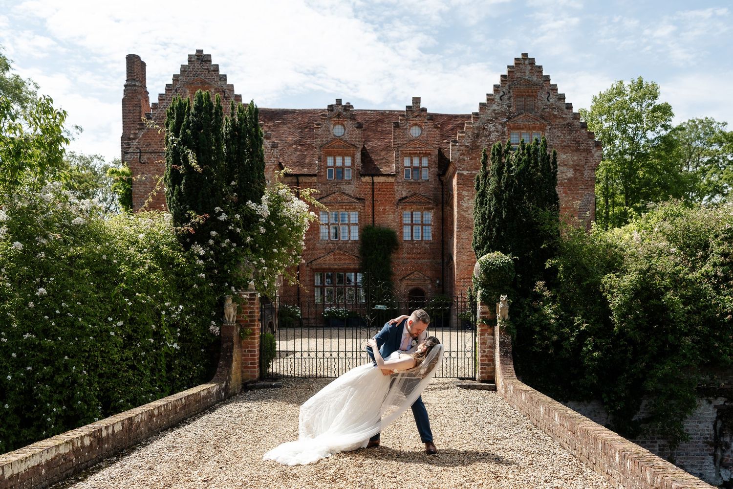 Bride and groom share romantic dip kiss on bridge before stunning historic red brick manor house and gardens.