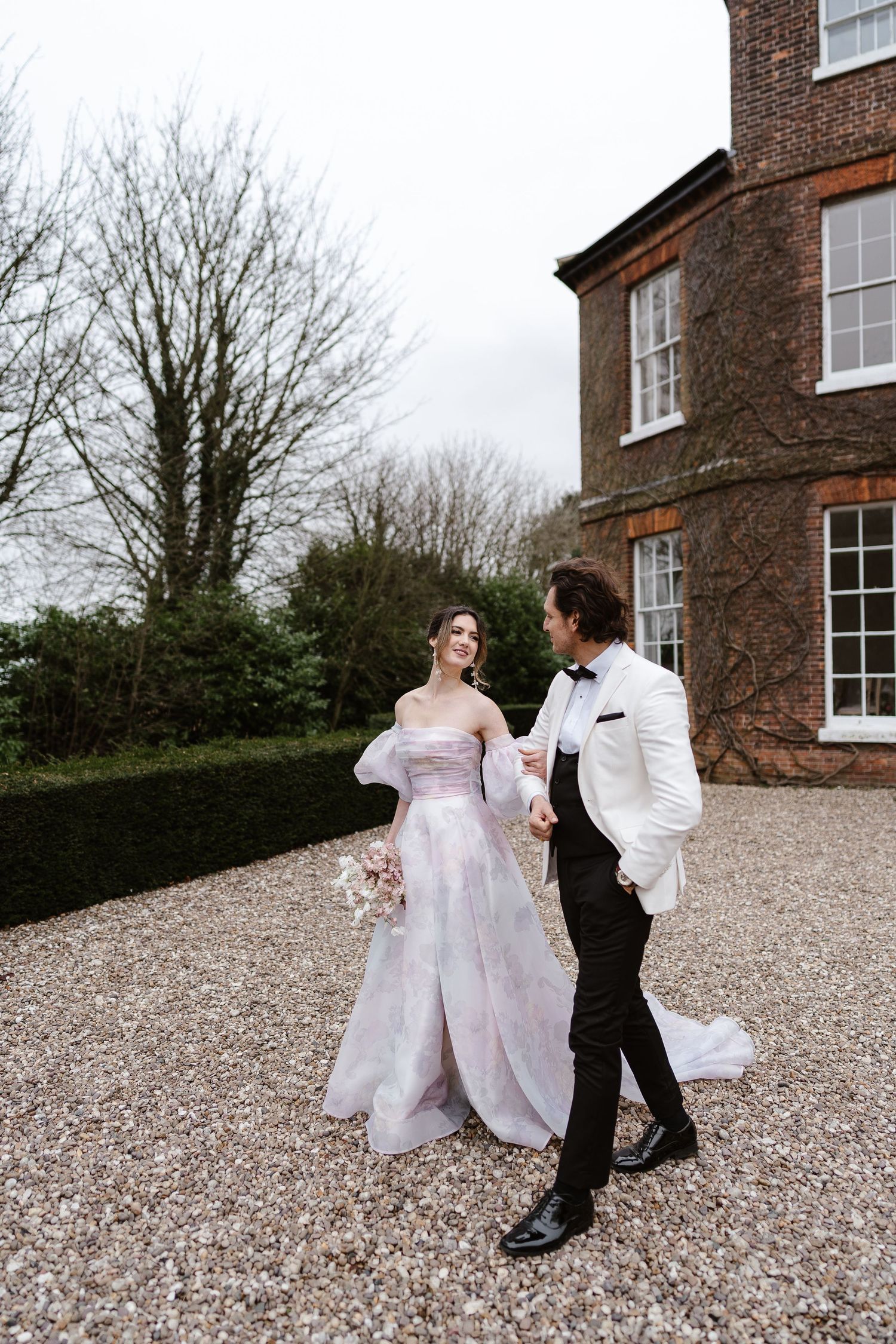 Bride in pink watercolor off-shoulder gown and groom in white tuxedo walking on gravel outside brick manor.