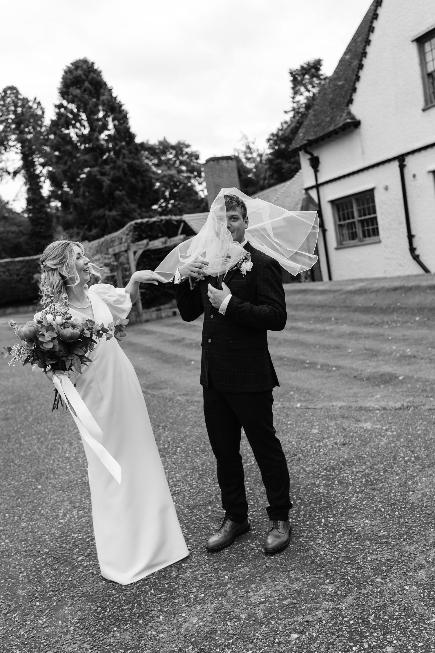 Bride laughs as veil blows over groom's face outside rustic white country house wedding venue.
