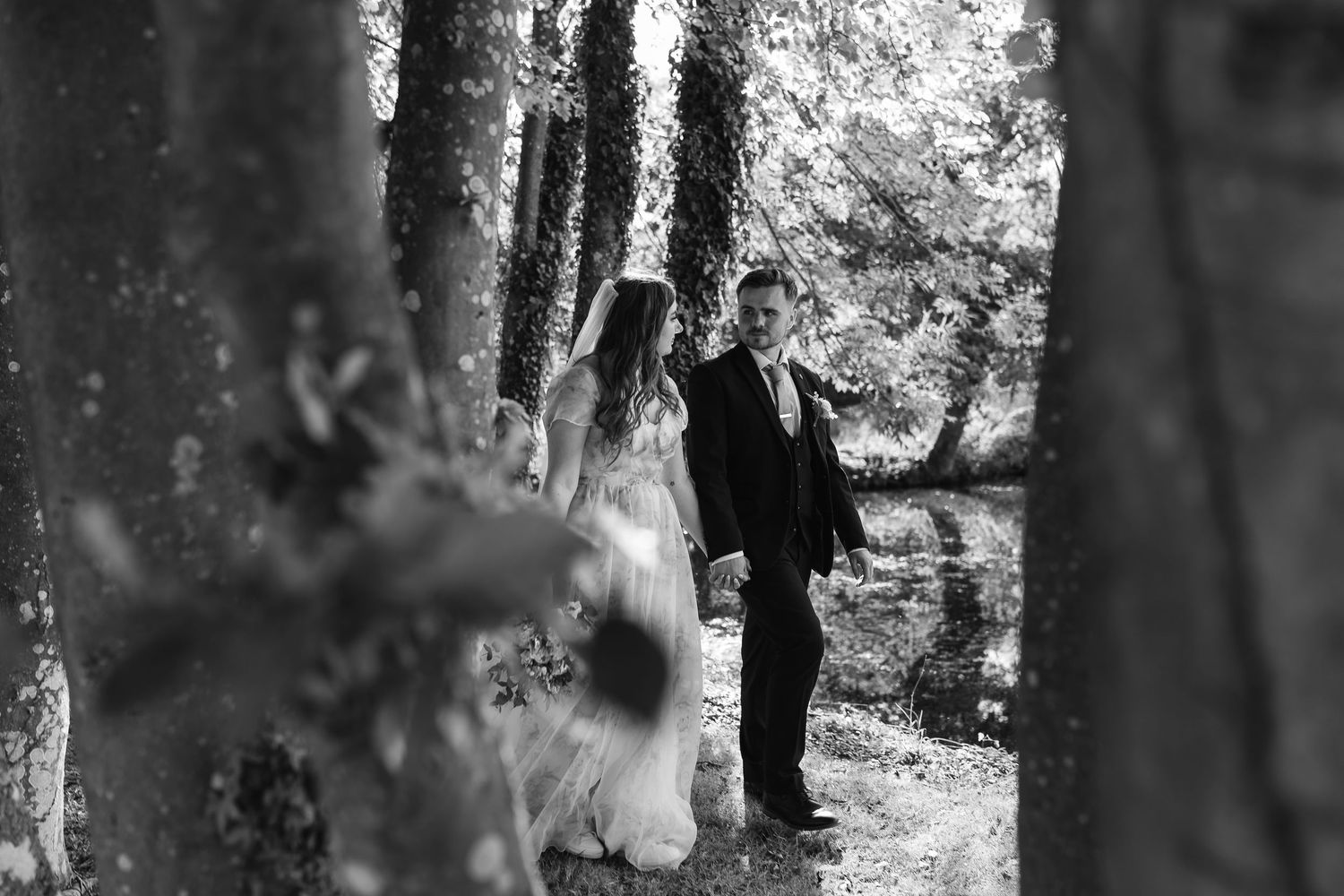 Black and white photo of bride and groom walking among tall trees at woodland wedding venue.