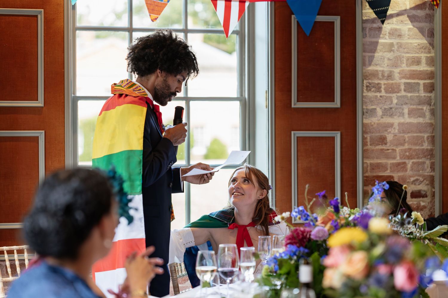 Man with rainbow flag gives speech at colorful wedding reception with floral centerpieces.