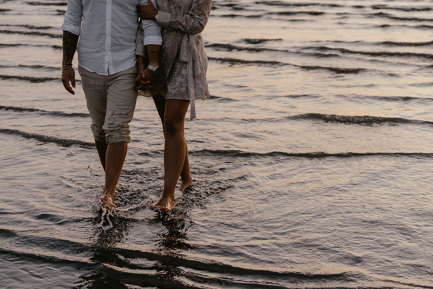 Barefoot couple walking together in shallow ocean water at dusk, close-up of legs.