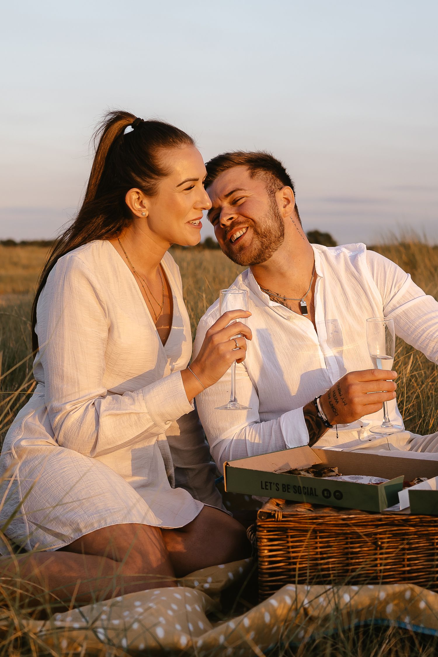Smiling couple laughing together at a picnic with champagne glasses and wicker basket in a field.