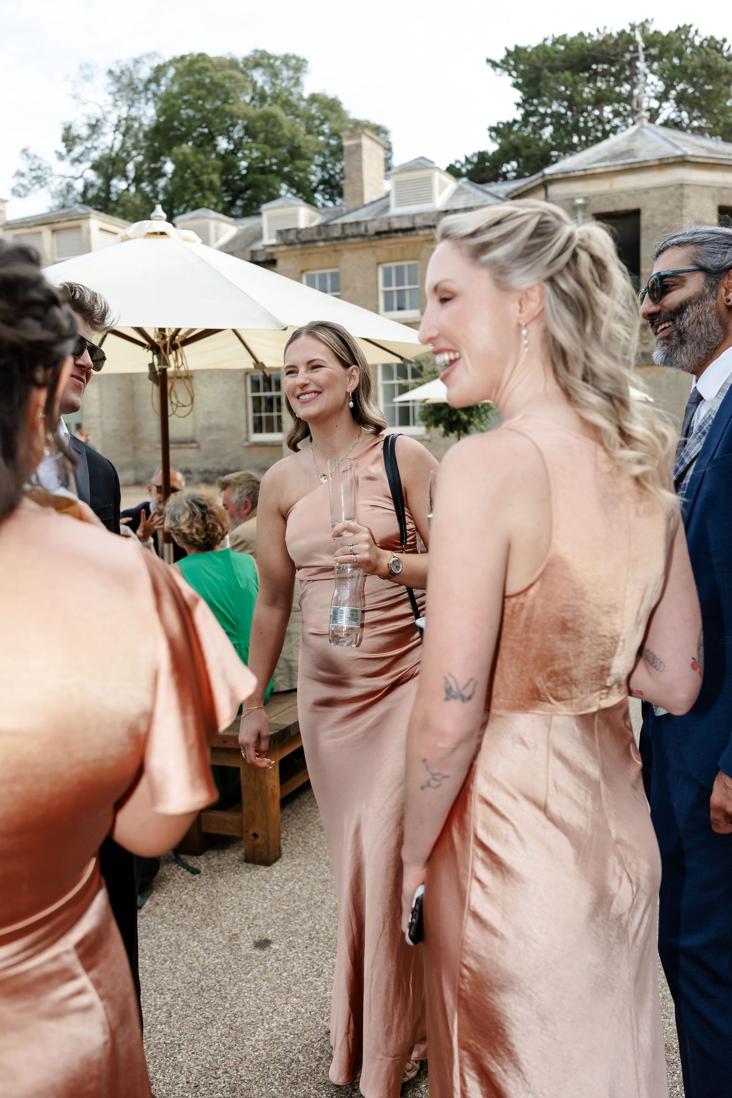 Bridesmaids in rose gold satin dresses laughing together at an outdoor wedding reception.