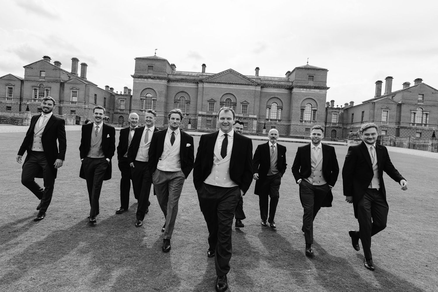 Groomsmen in morning suits walk confidently together outside a grand English country house in black and white.
