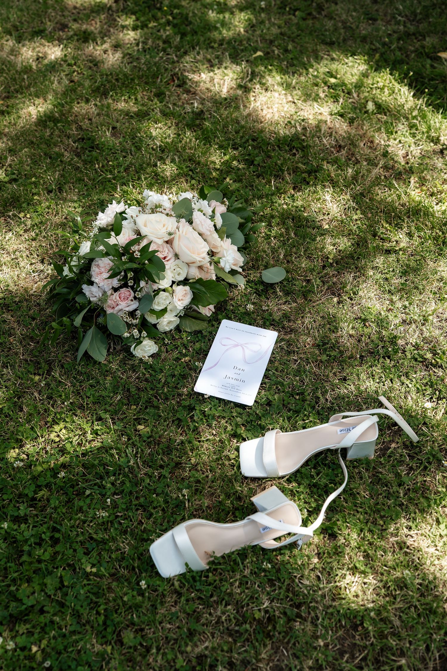 Bridal bouquet, wedding invitation, and white heeled sandals arranged on green grass in natural sunlight.