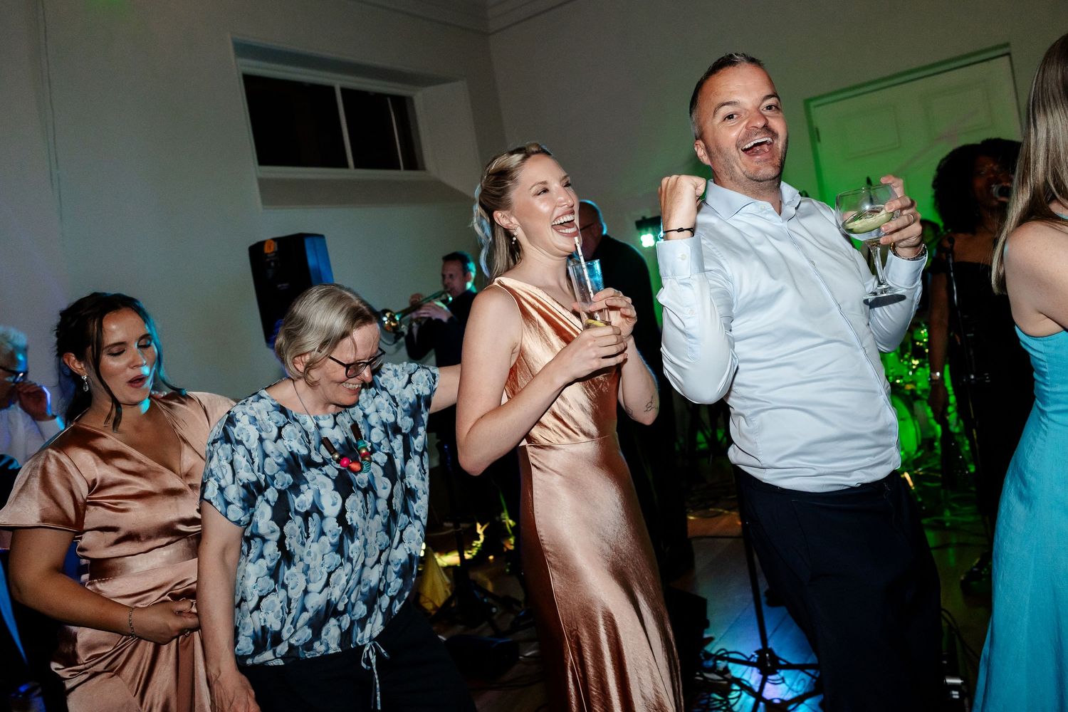 Bridesmaid in gold satin dress sings into microphone while dancing with guest at wedding party.