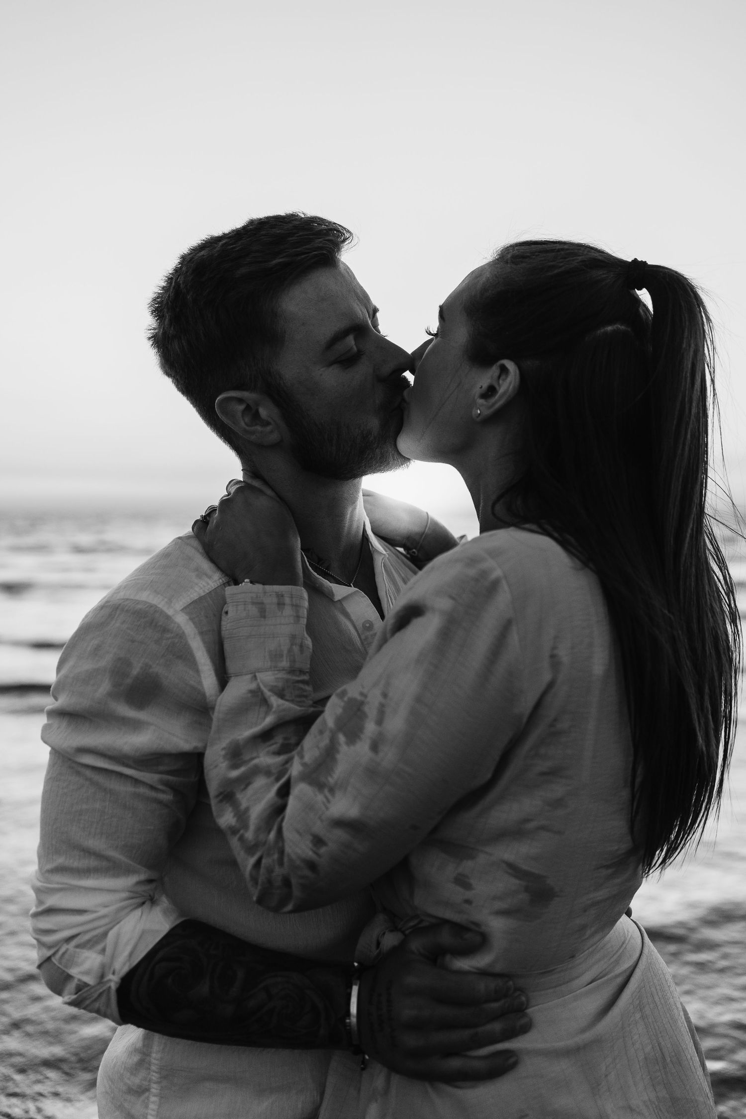 Black and white close-up of couple sharing a kiss on beach at sunset.