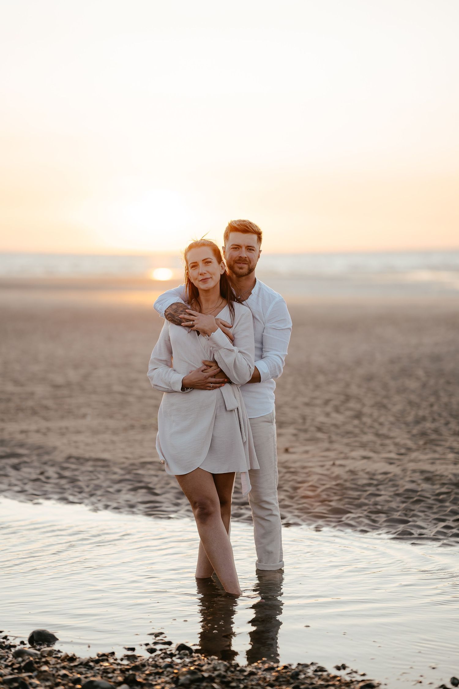 A loving couple standing barefoot in shallow beach water, embracing during a warm golden sunset.