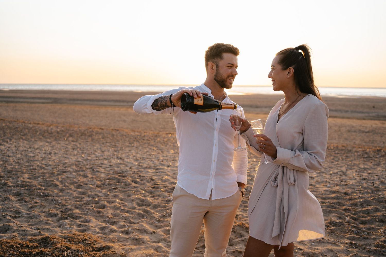 Smiling couple on a sunny beach, man pouring Canti champagne into a glass for his partner.