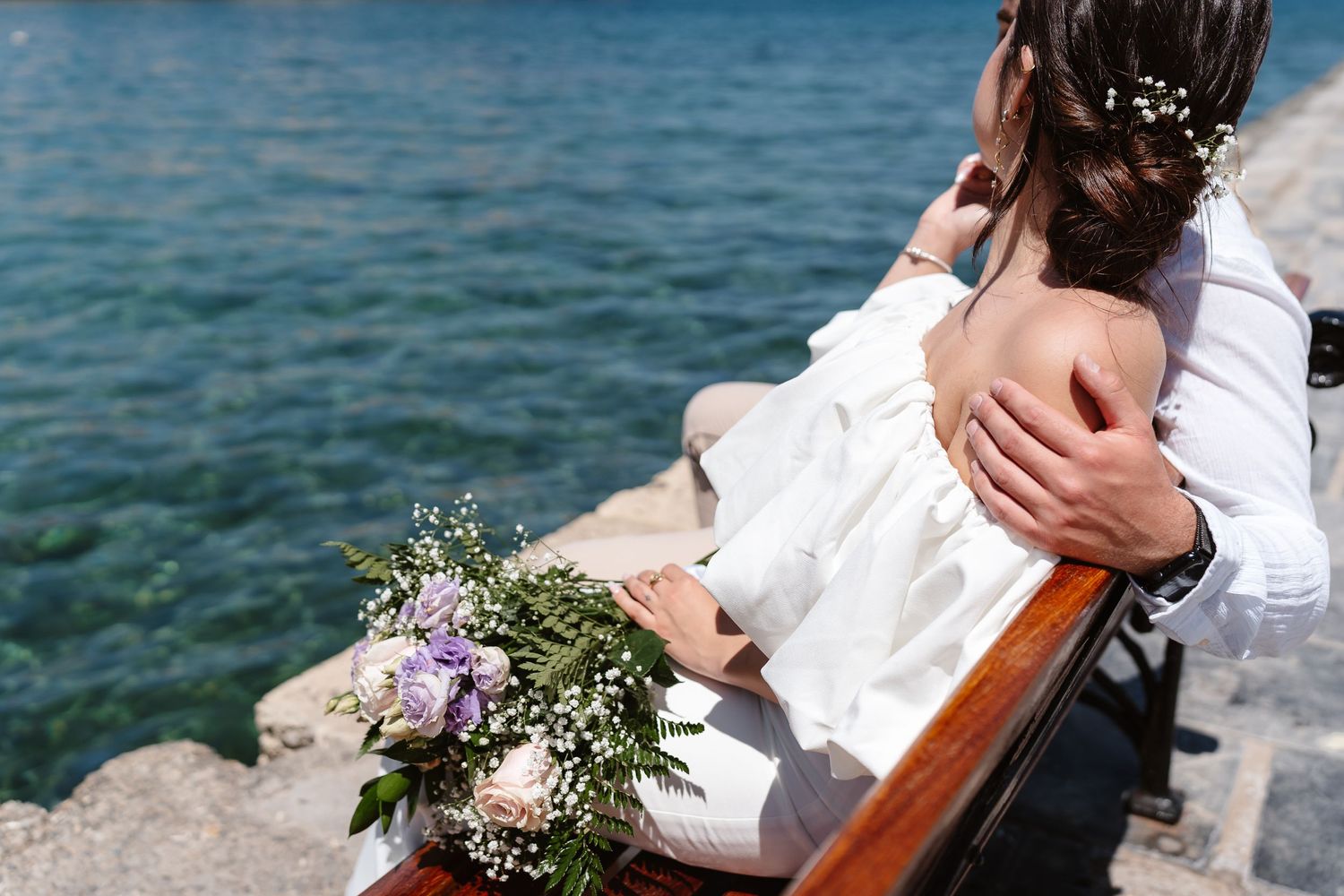 Bride and groom sitting on a waterfront bench embracing with a bouquet overlooking turquoise sea water.