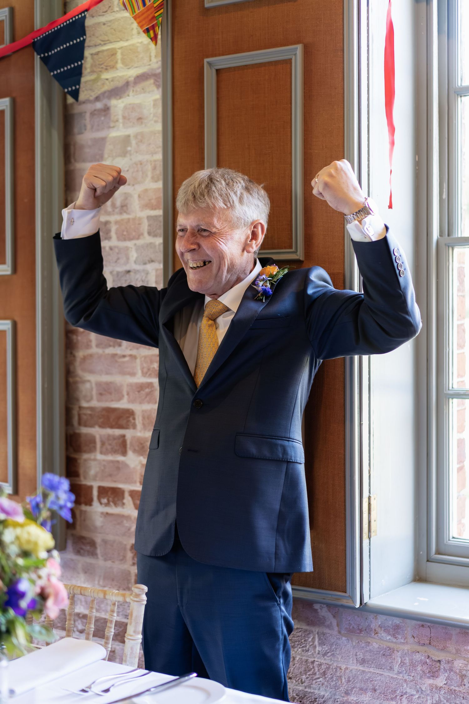 Jubilant older man in navy suit with gold tie raises both fists in celebration at a wedding reception.