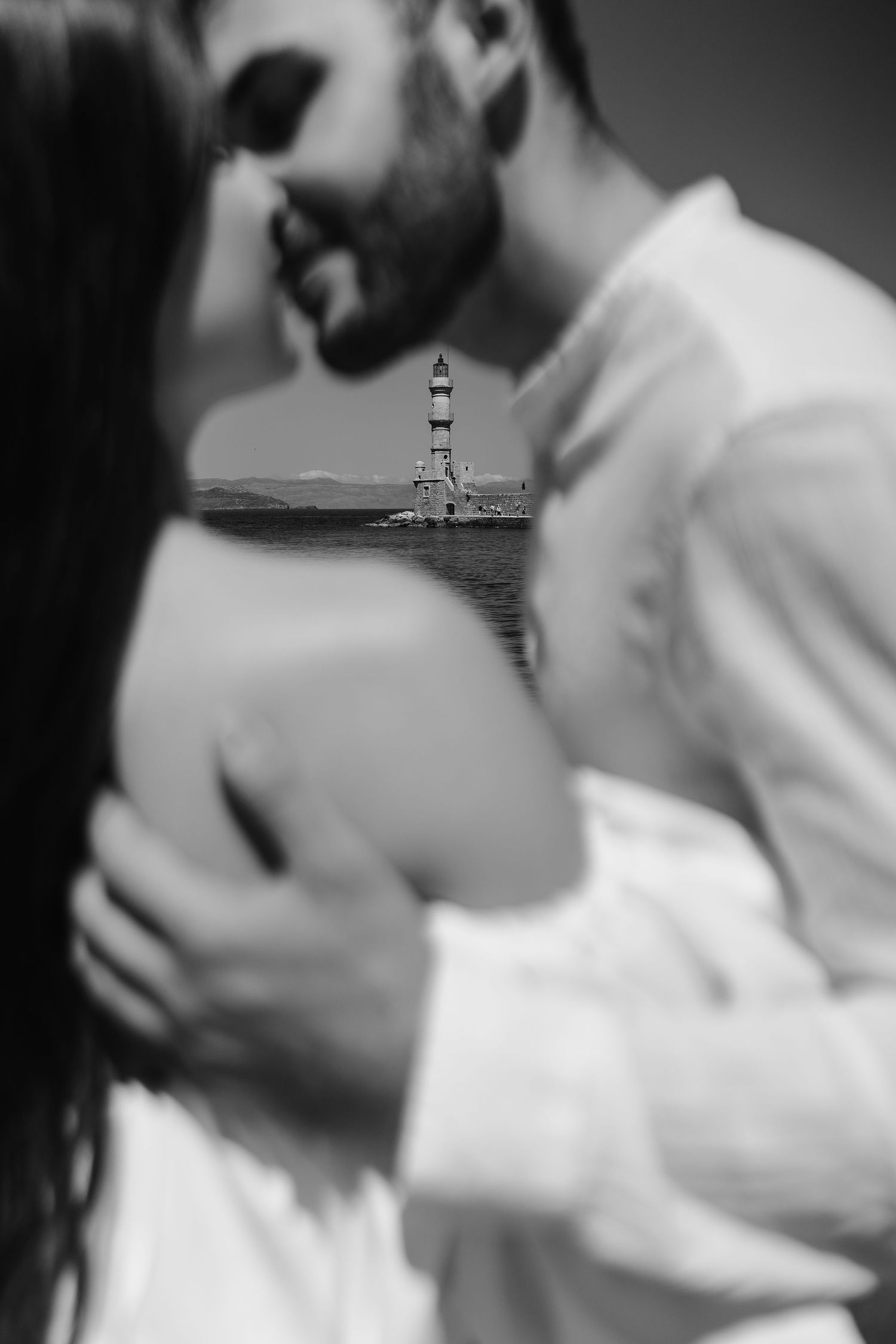Black and white photo of newlyweds embracing with Chania lighthouse between them in Greece.
