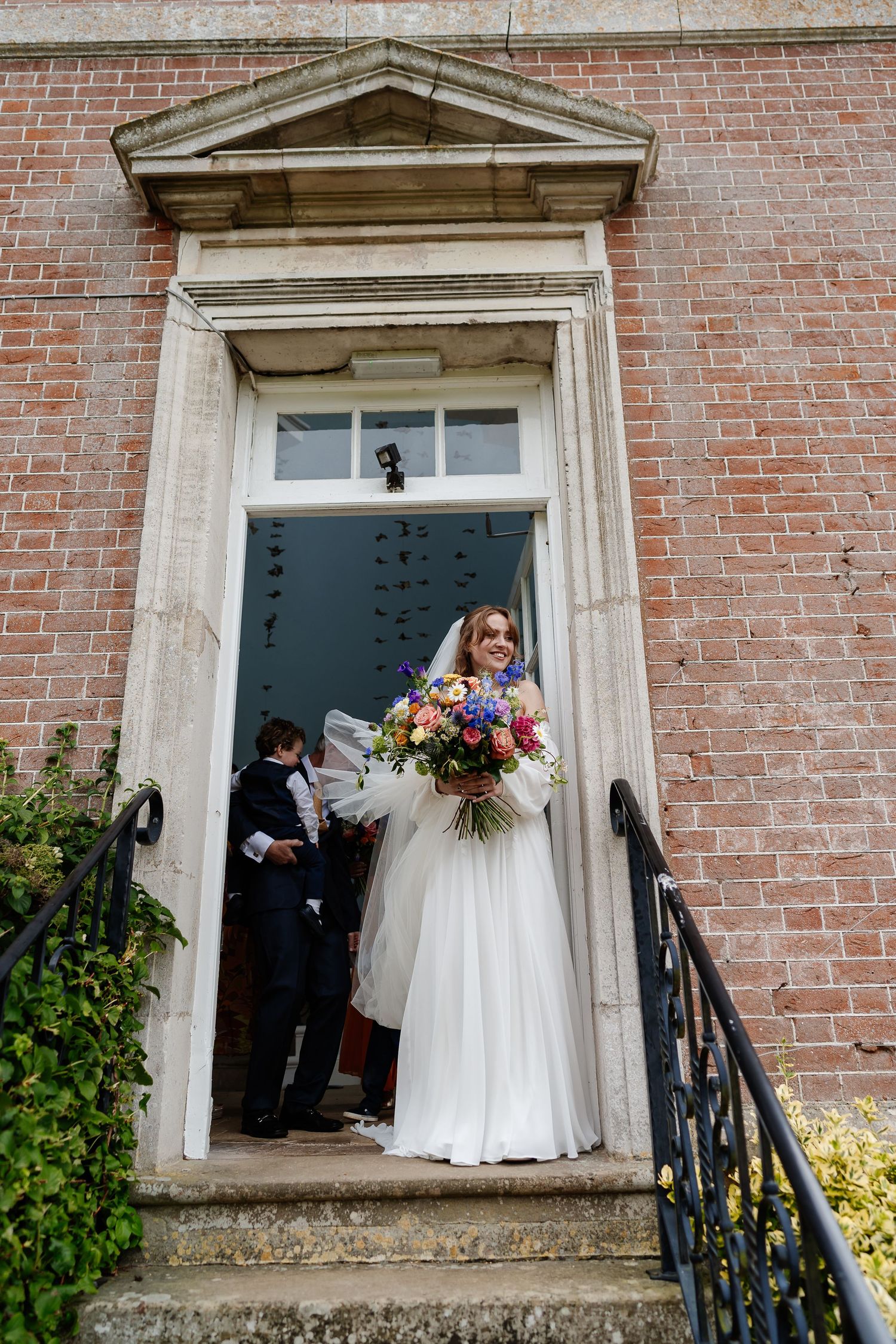Bride in white gown holding colorful bouquet standing in doorway of historic red brick wedding venue.