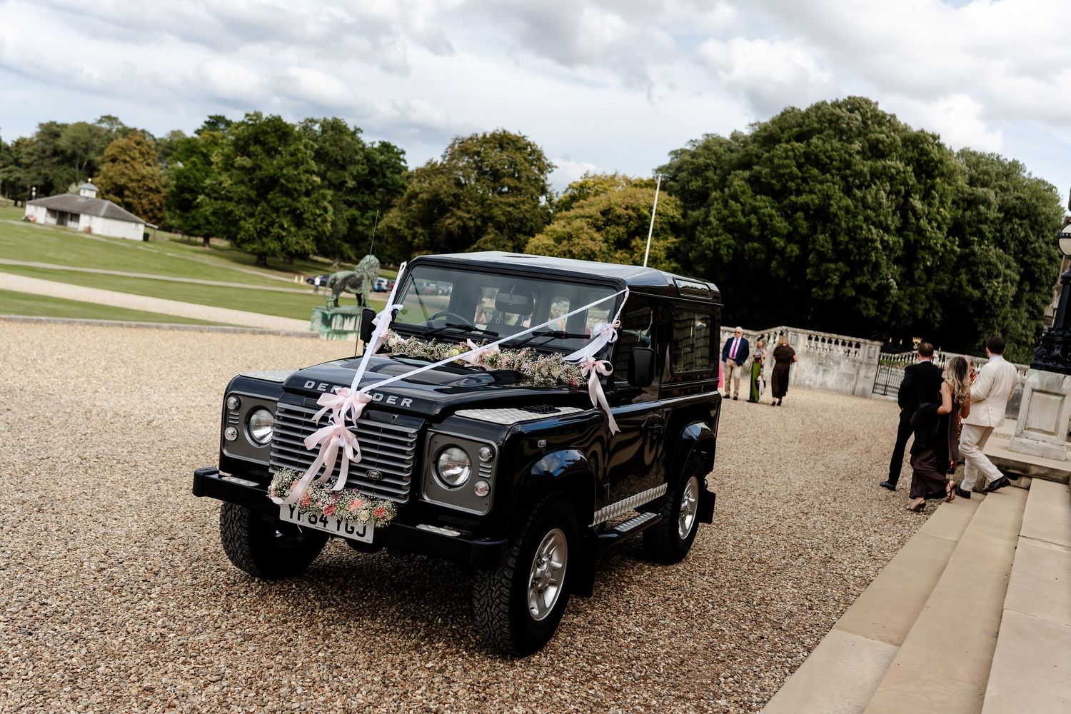 Black Land Rover Defender wedding car decorated with white ribbons and flowers at a countryside venue.