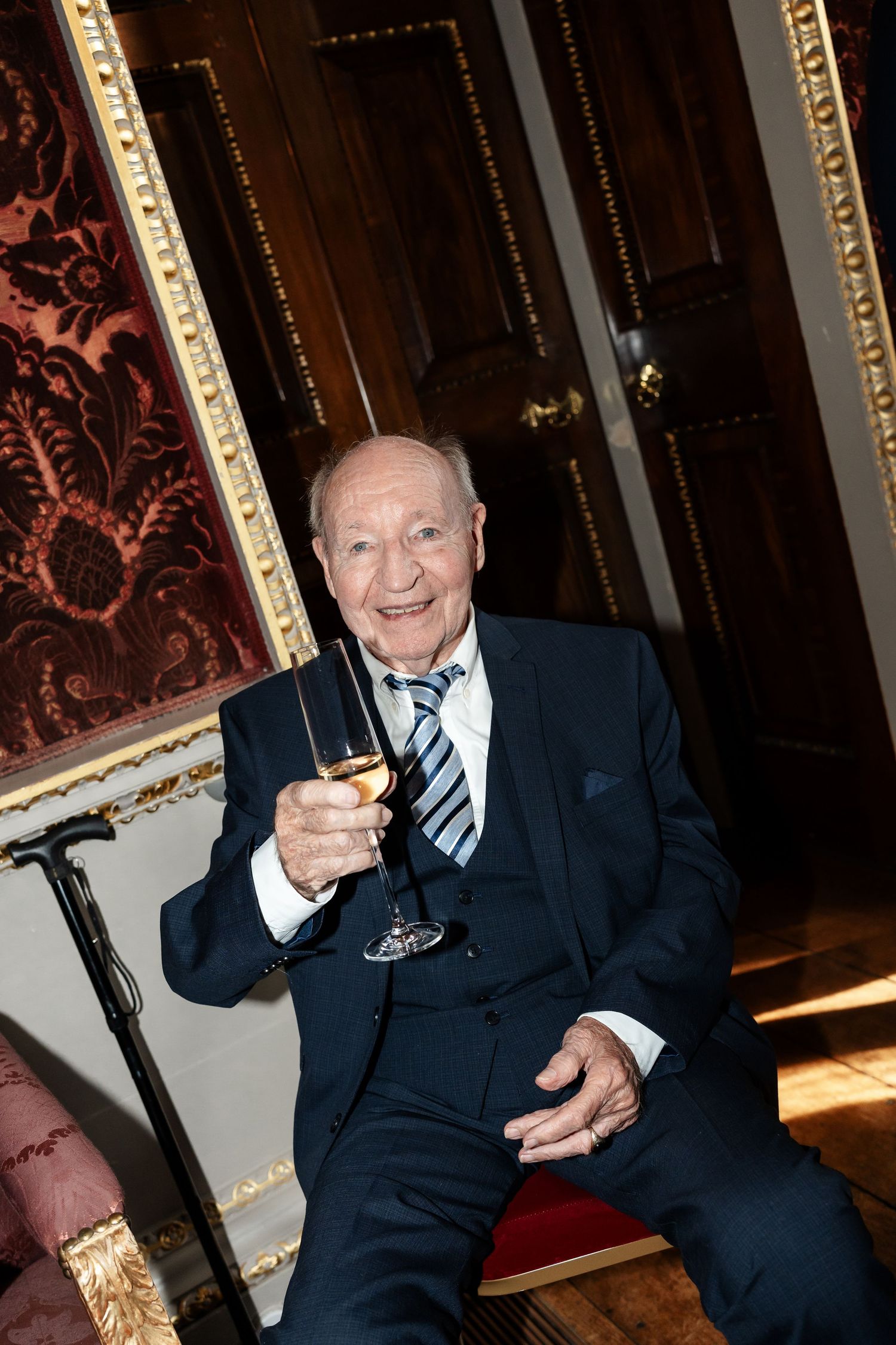 Elderly gentleman in navy suit raises champagne glass in celebratory toast at luxurious wedding reception.