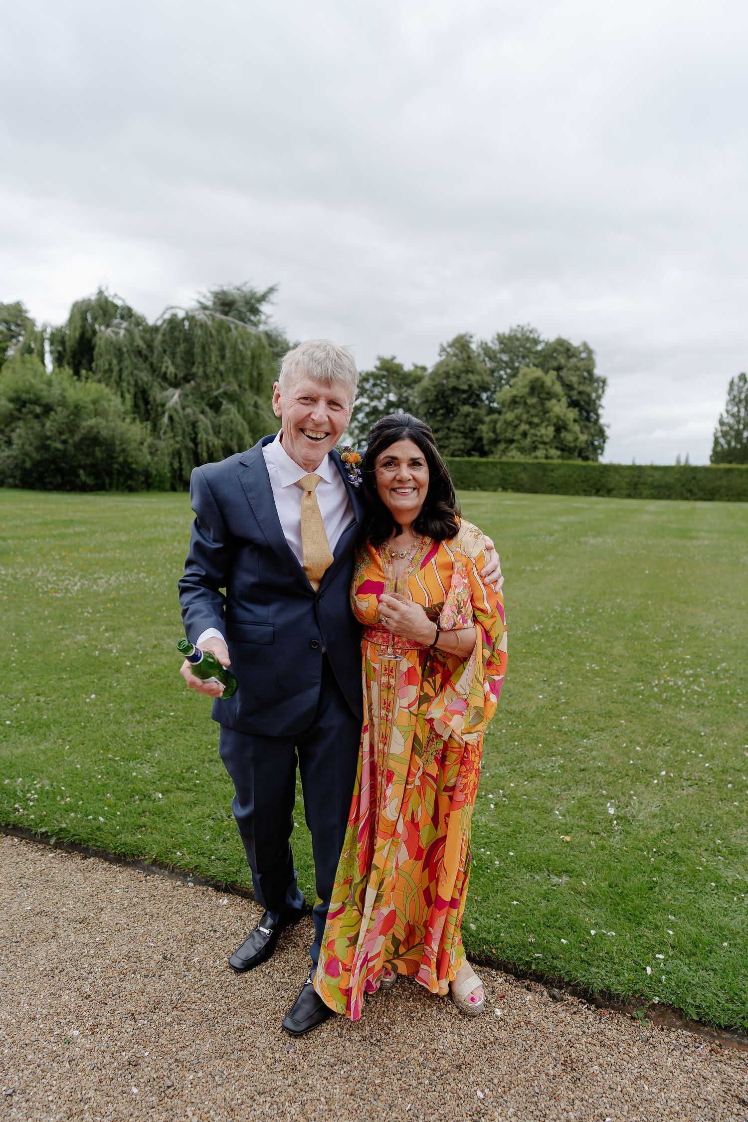 Smiling couple in formal wedding guest attire pose together on gravel path in manicured garden.