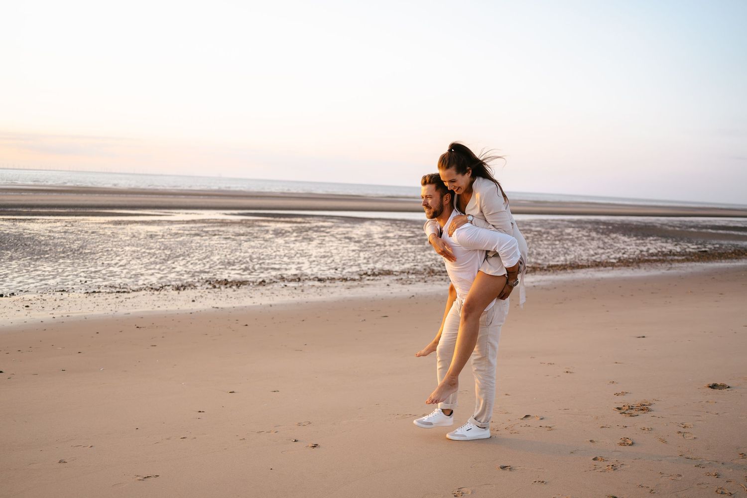 Man giving woman a piggyback ride on beach at sunset near ocean waves.