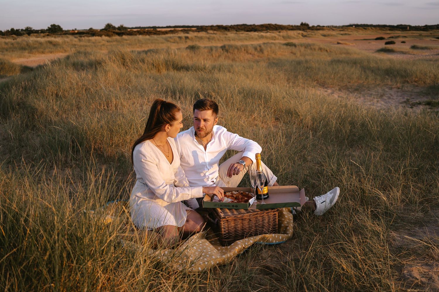 Couple enjoying a romantic picnic with champagne in a golden grass field at sunset.