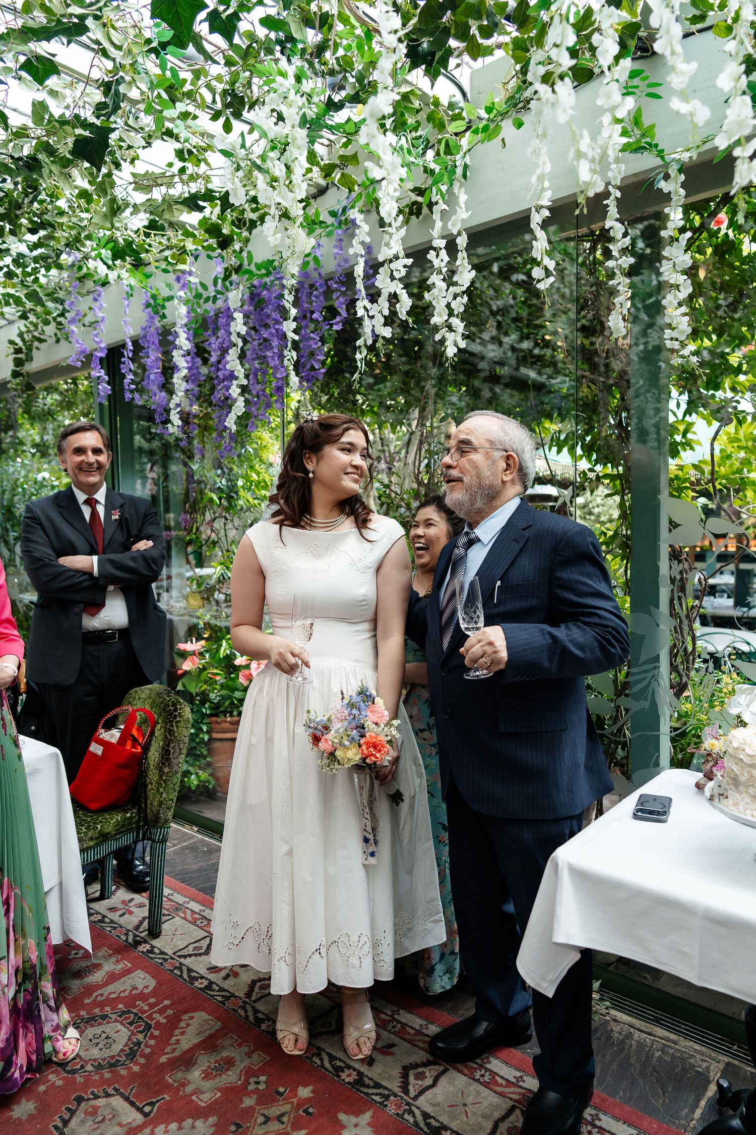 Bride holding colorful bouquet stands with older gentleman under wisteria in a garden venue.
