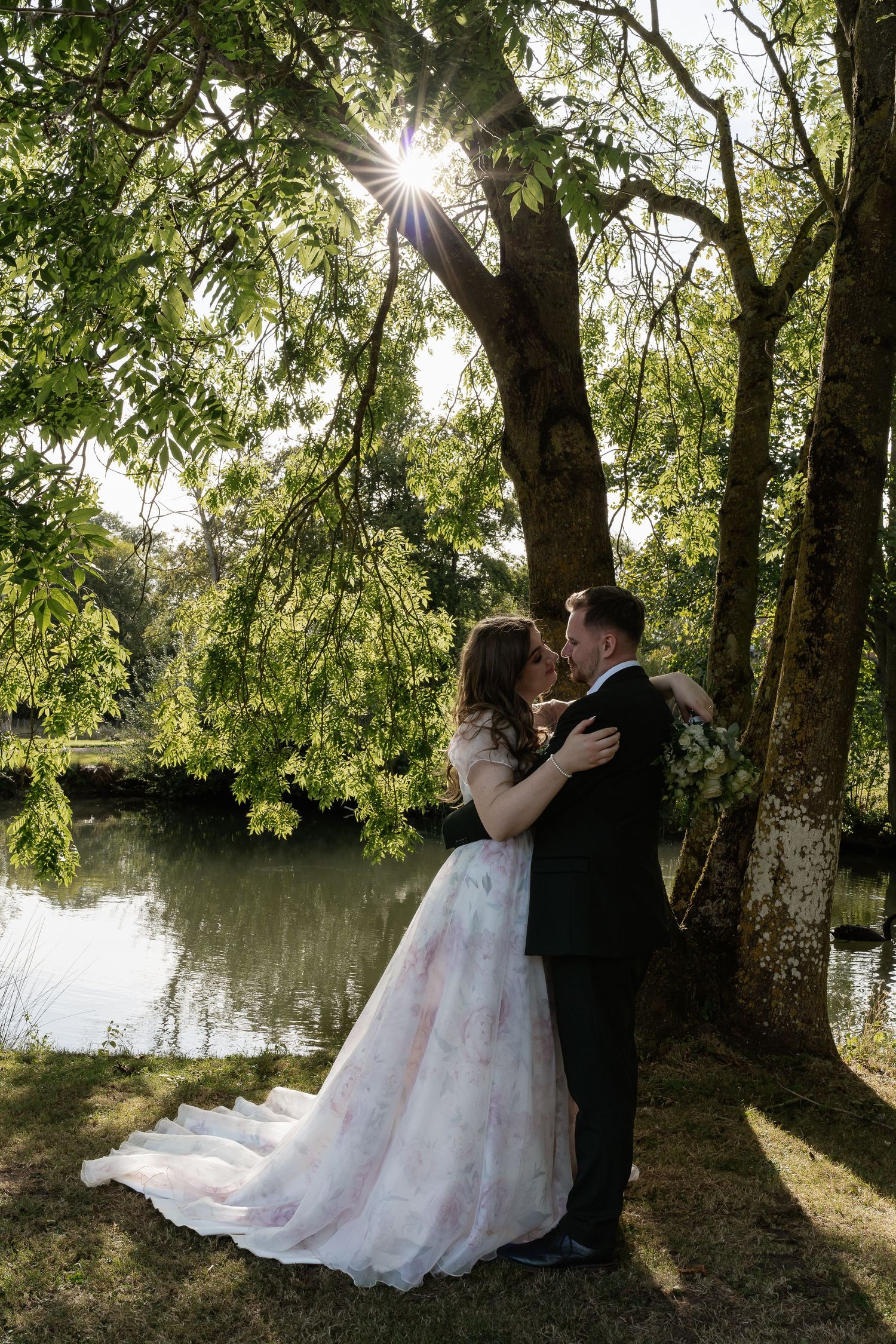 Bride and groom share romantic kiss under sunlit tree by tranquil pond in lush garden setting.
