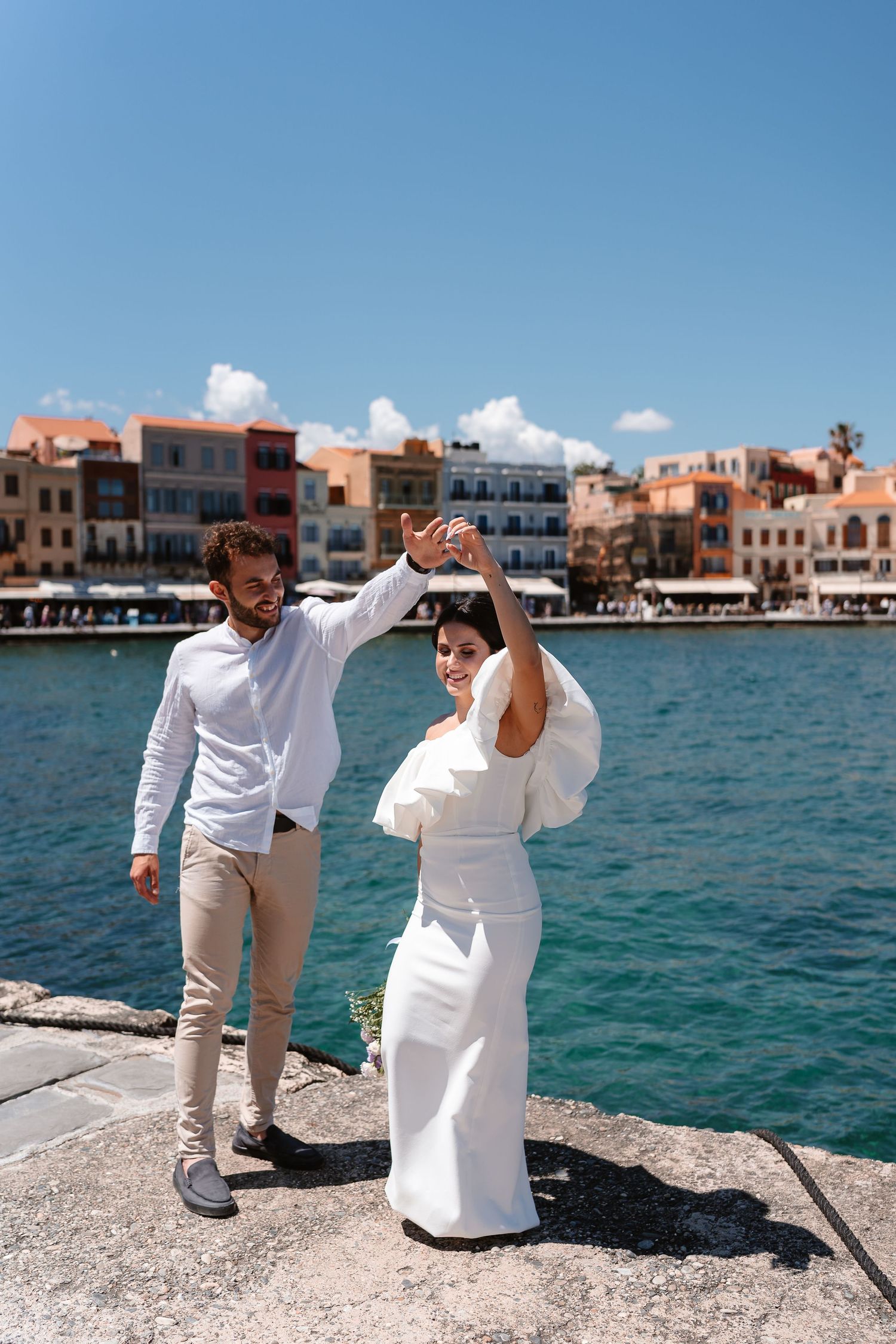 Groom twirls bride in white wedding dress on a sunny Greek harbor pier with vibrant waterfront buildings behind them.
