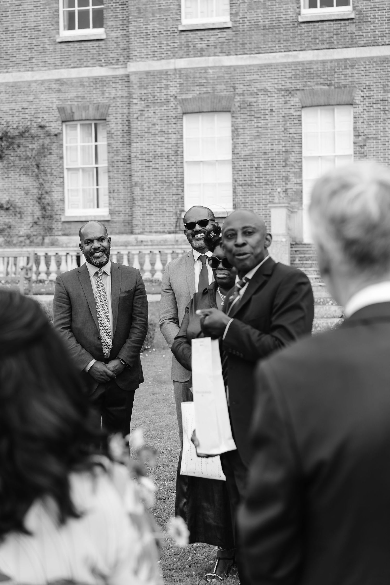 Black and white photo of man reading from paper during outdoor wedding ceremony at brick manor house.