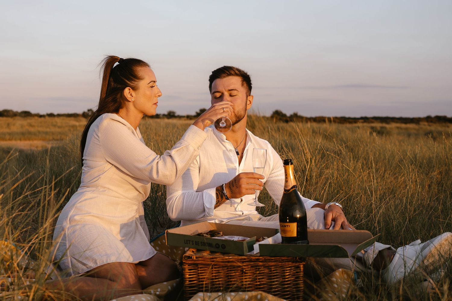 Woman feeding man during a romantic sunset picnic in a grassy field with champagne and pizza.