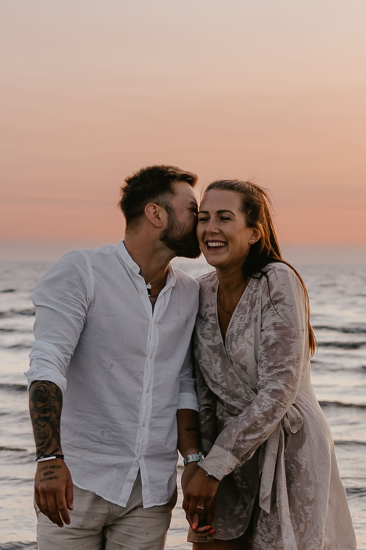 Man kissing woman on cheek at sunset beach, she laughs joyfully while holding hands.
