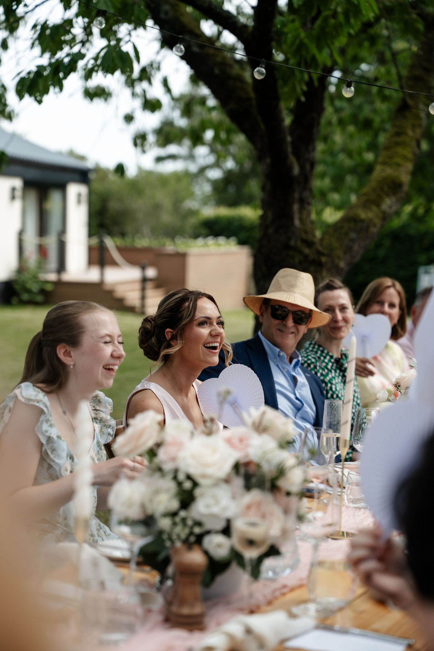 A laughing bride sits among smiling guests at a beautifully decorated outdoor wedding reception table.