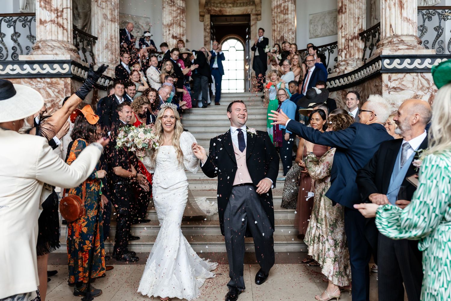 A bride and groom celebrate with confetti as guests cheer on grand ornate stone steps.