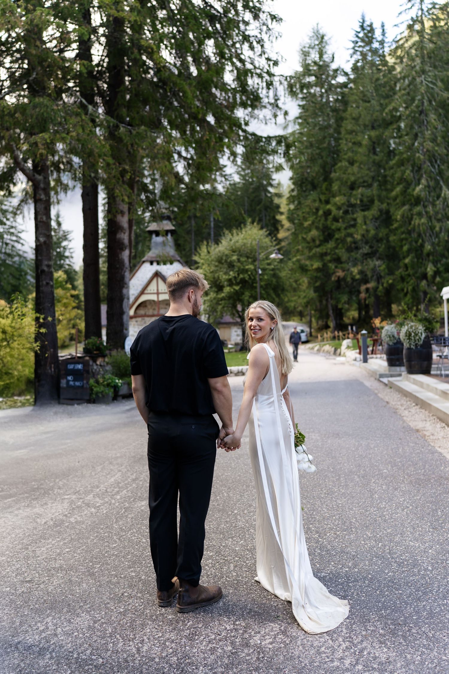 Bride in elegant white gown looks back smiling while holding grooms hand on a tree-lined road.