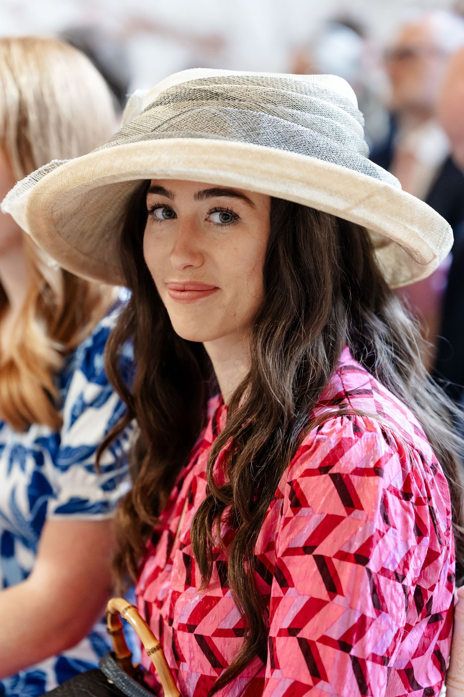Young woman with long dark hair wearing a wide-brim straw hat and pink geometric print dress at outdoor event.