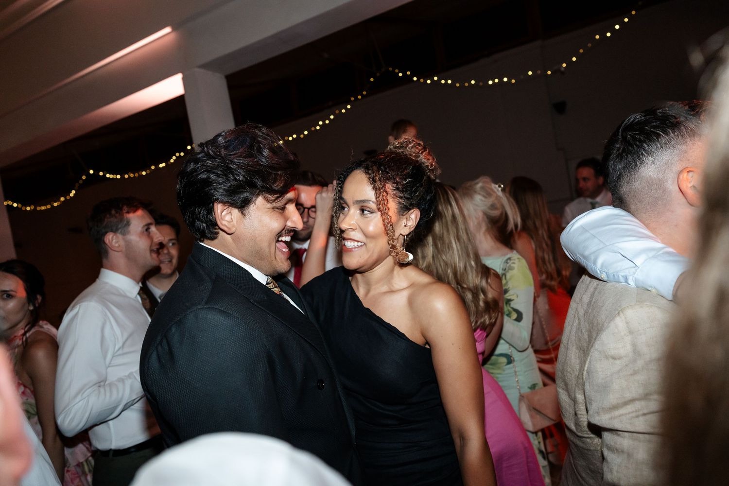 Couple smiling and dancing together at a wedding reception with fairy lights and guests in the background.