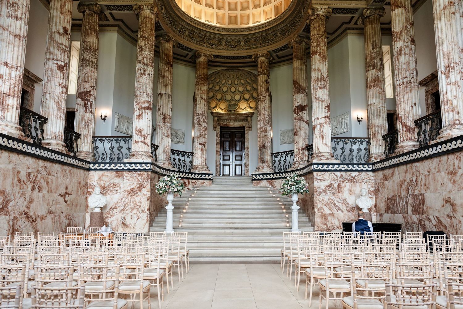 Elegant wedding ceremony setup in a grand neoclassical hall with marble columns, chiavari chairs, and floral arrangements.