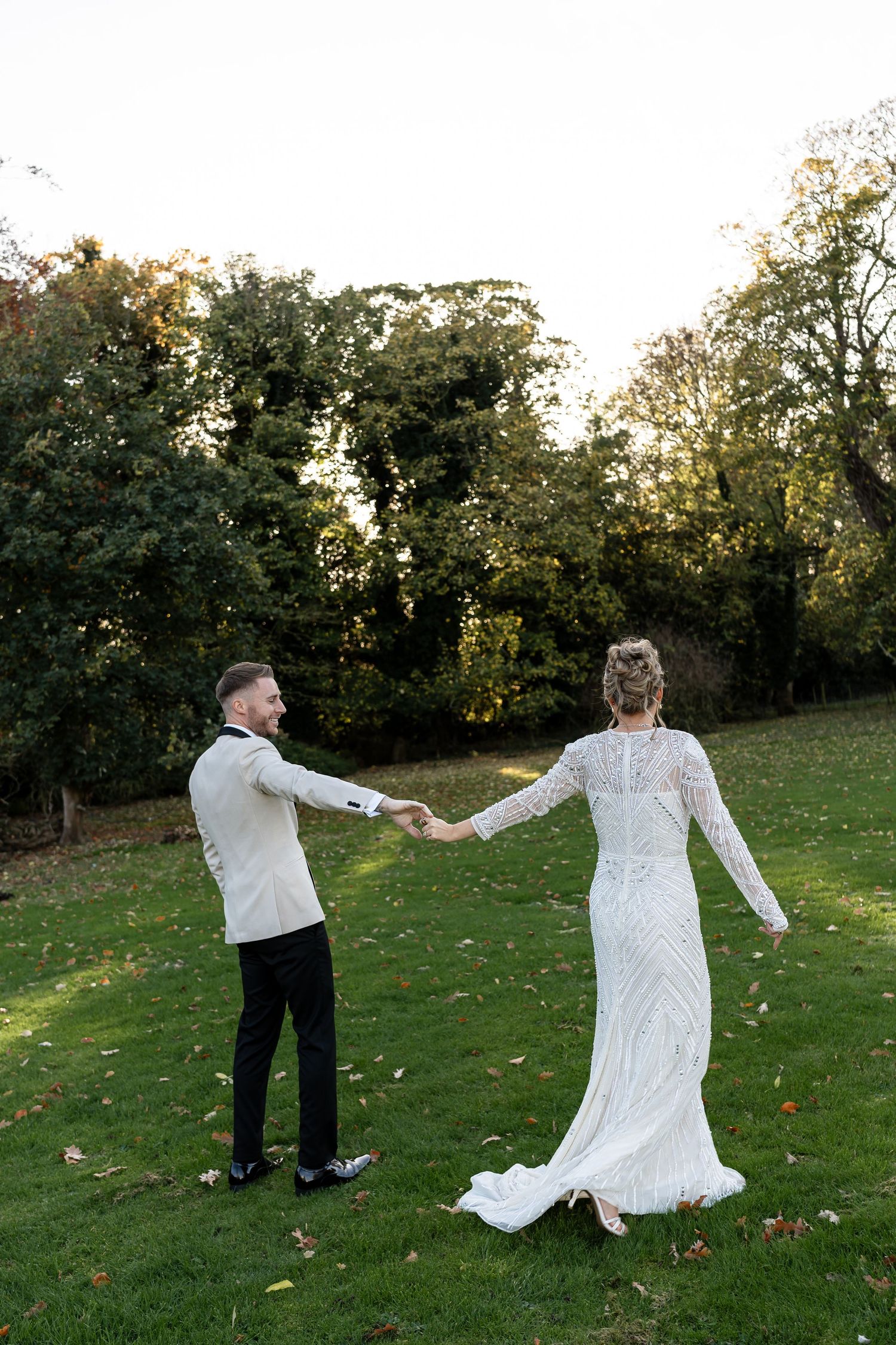 Bride and groom holding hands and dancing on a green lawn surrounded by autumn trees.