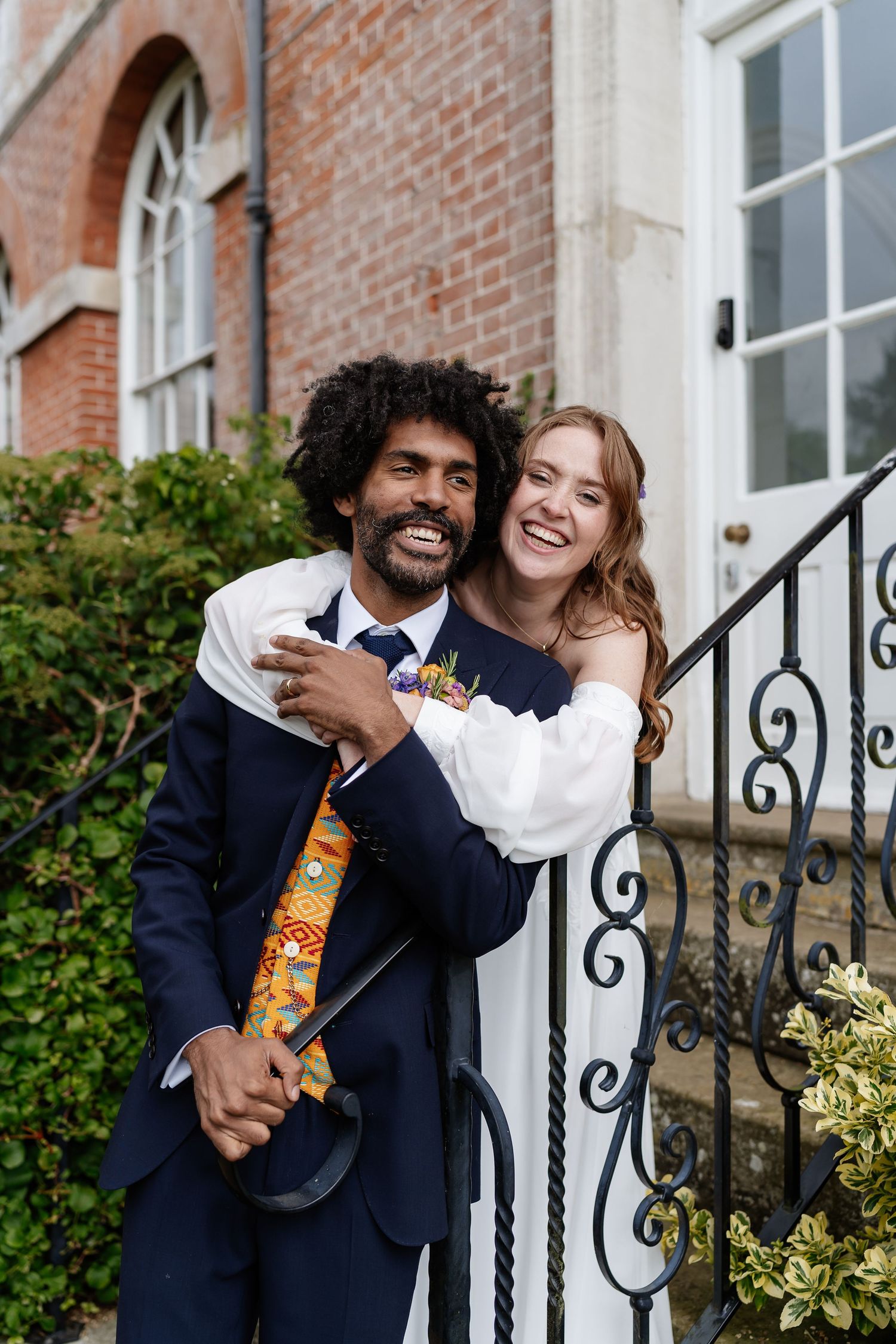 Joyful interracial couple embracing on steps outside a brick building on their wedding day.