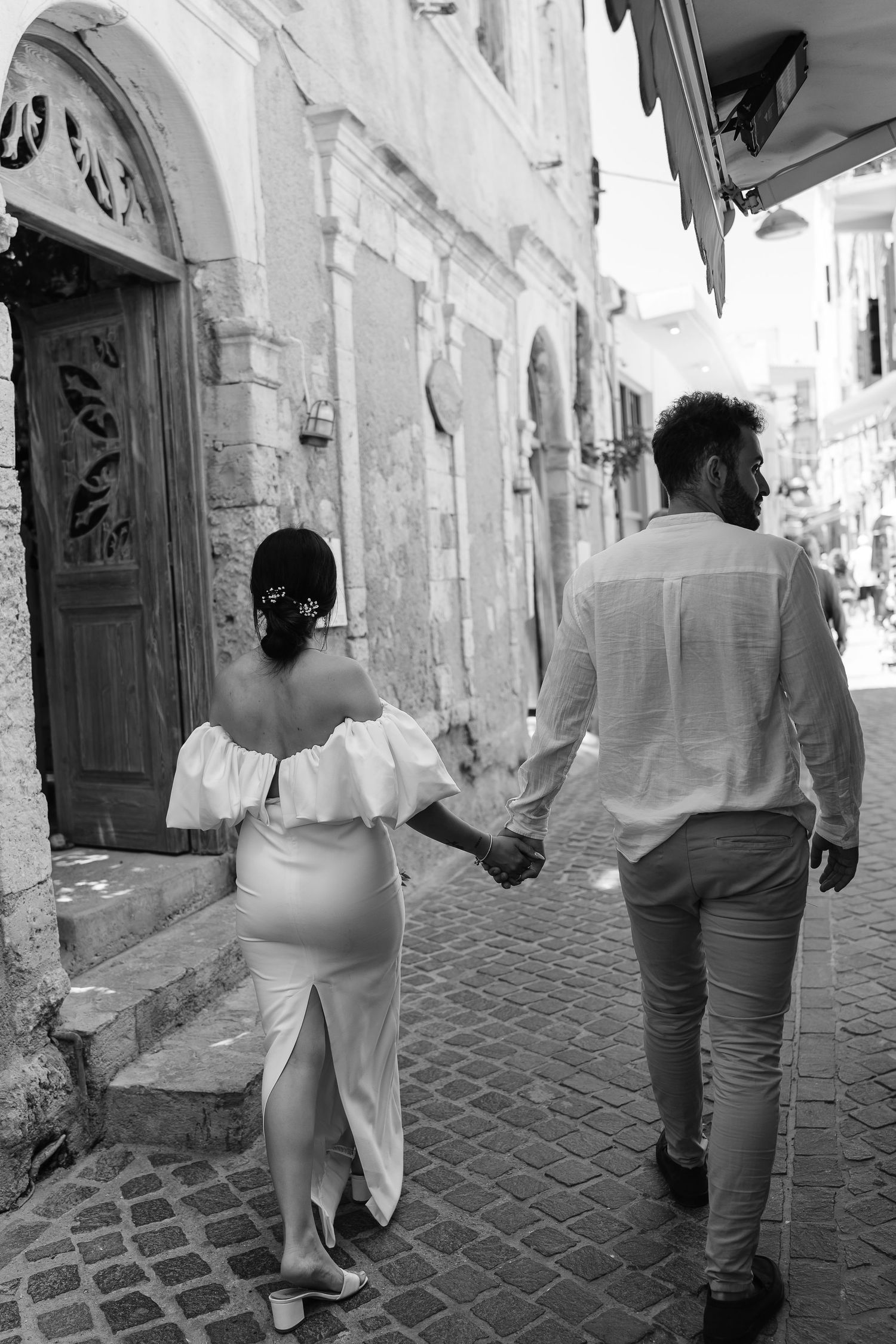 Black and white photo of newlyweds holding hands walking through charming cobblestone Mediterranean street.