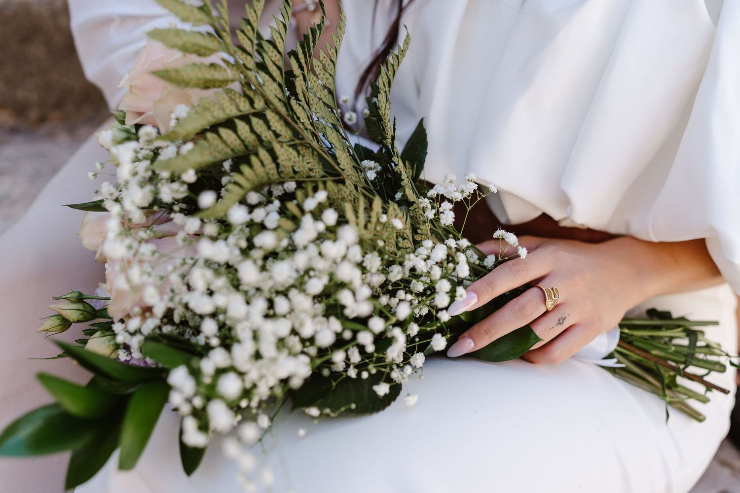 Bride holding elegant wedding bouquet of white baby's breath, roses, and green ferns in white dress.